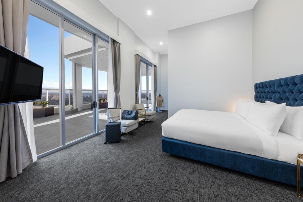 Meriton Suites Bondi Junction Сидней Экстерьер фото The photo shows a modern and spacious hotel room. It features a large bed with a blue headboard, a flat-screen TV mounted on the wall, and a cozy seating area with a chair and small table. The room has large windows that offer natural light and a vie