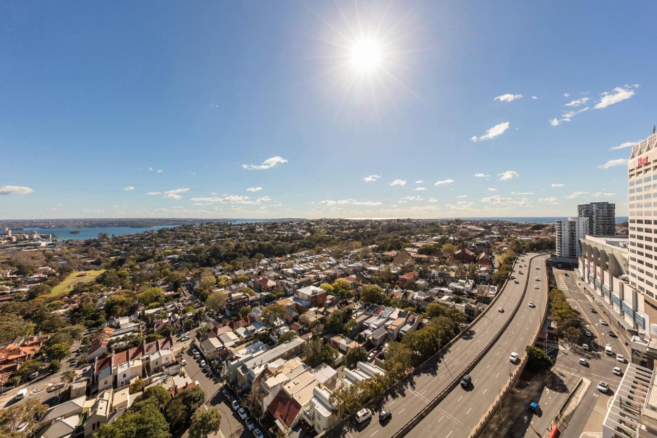 Meriton Suites Bondi Junction Сидней Экстерьер фото The photo shows a panoramic view of a city landscape under a clear blue sky. In the foreground, there is a road with vehicles traveling along it, bordered by trees and buildings. In the background, you can see a residential area with various houses, 