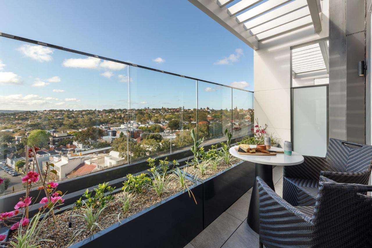 Meriton Suites Bondi Junction Сидней Экстерьер фото The photo shows a modern balcony with a glass railing, enhancing the view of the surrounding area, which includes various rooftops and greenery in the distance. There is a small round table set with a decorative floral arrangement and some items, lik