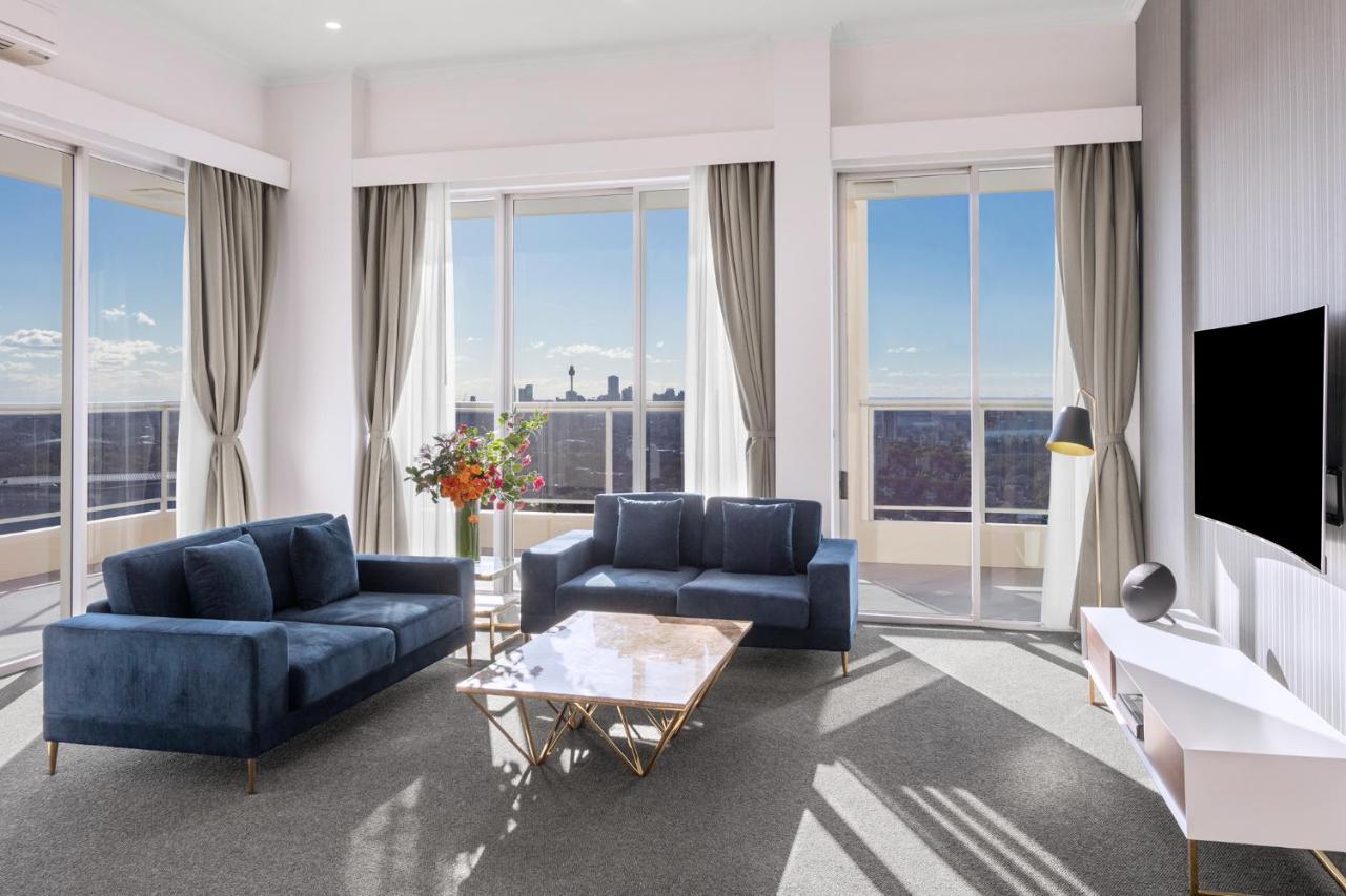 Meriton Suites Bondi Junction Сидней Экстерьер фото The photo shows a modern living room with large windows that allow natural light to fill the space. There are two blue sofas arranged around a coffee table with a glass top and gold legs. A floral arrangement adds a touch of color, placed on a nearby