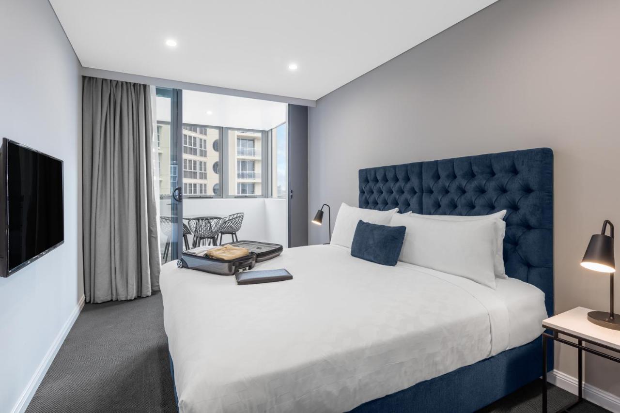 Meriton Suites Bondi Junction Сидней Экстерьер фото The photo shows a modern hotel room featuring a large bed with a tufted blue headboard. There are minimalistic bedside tables with lamps on either side of the bed. A flat-screen TV is mounted on the wall across from the bed. To one side, there are fl