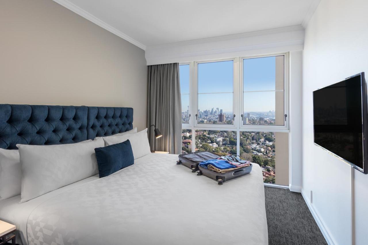 Meriton Suites Bondi Junction Сидней Экстерьер фото The photo shows a modern hotel bedroom featuring a king-sized bed with a blue tufted headboard. The bed is neatly made with white linens and a decorative blue pillow. There is a large window that offers a view of the city skyline, allowing natural li