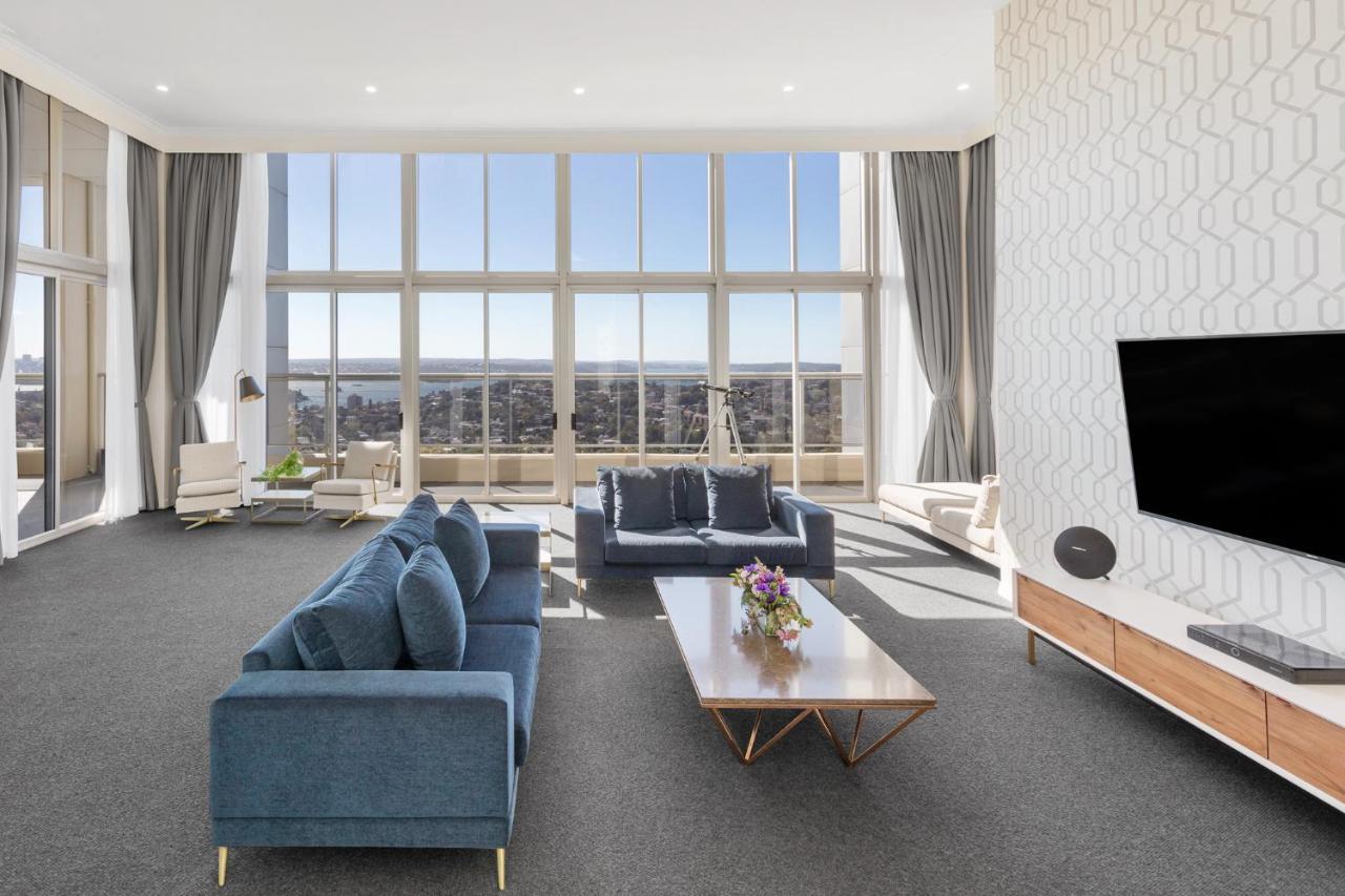 Meriton Suites Bondi Junction Сидней Экстерьер фото The photo shows a modern and spacious living area. It features large windows that provide a panoramic view of the outside landscape and allow natural light to fill the room. The interior design includes a blue sectional sofa placed in the center, com