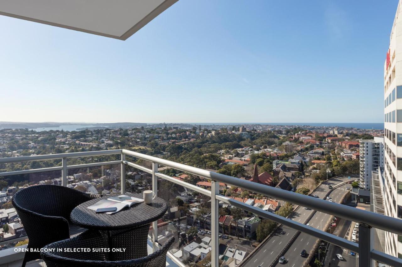 Meriton Suites Bondi Junction Сидней Экстерьер фото The photo shows a balcony with a view over a cityscape that includes buildings and greenery, extending towards the horizon where the sea is visible. The balcony features a small round table and two black chairs, suggesting a space for relaxation or d