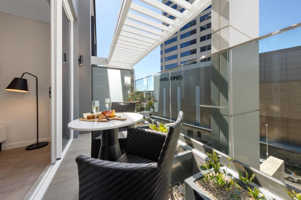 Meriton Suites Bondi Junction Сидней Экстерьер фото The photo shows a modern balcony area with glass railings. It features a small round table set for two, with a plate of food and drinks. There are two stylish wicker chairs positioned around the table. The space is bright and airy, with sunlight stre