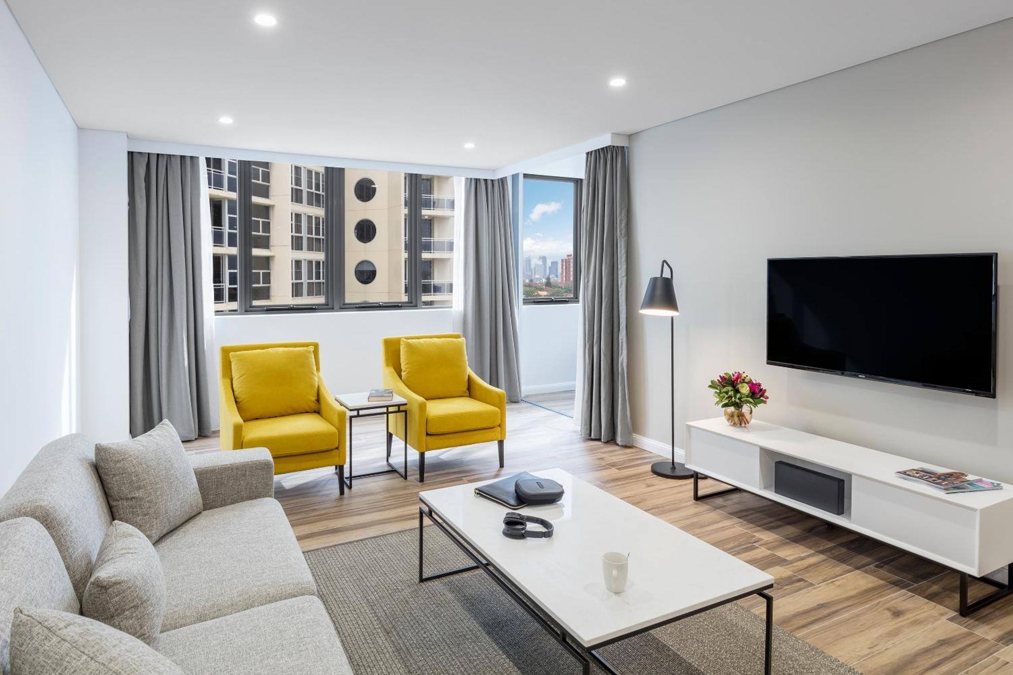 Meriton Suites Bondi Junction Сидней Экстерьер фото The photo shows a modern, well-lit living room. It features a light gray sofa with a textured fabric. There are two bright yellow armchairs positioned near a window, which allows natural light to enter the space. The room has light gray curtains and 